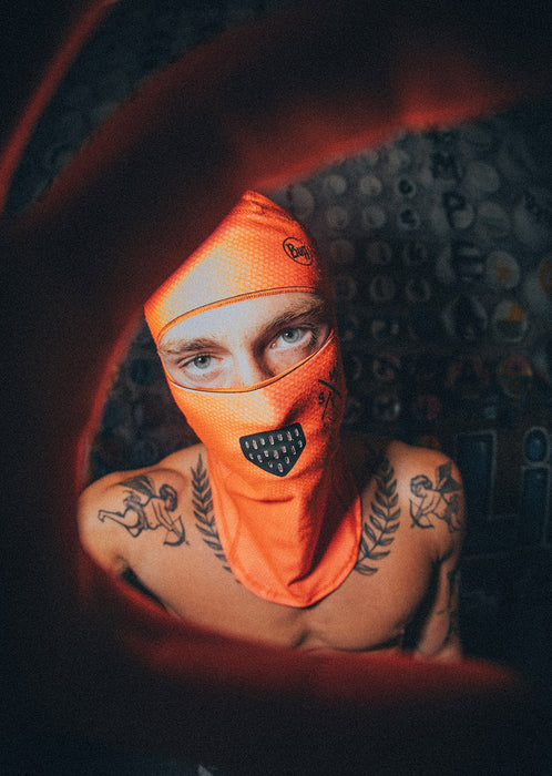 Person wearing an orange balaclava with a brand logo, standing against a dark background.