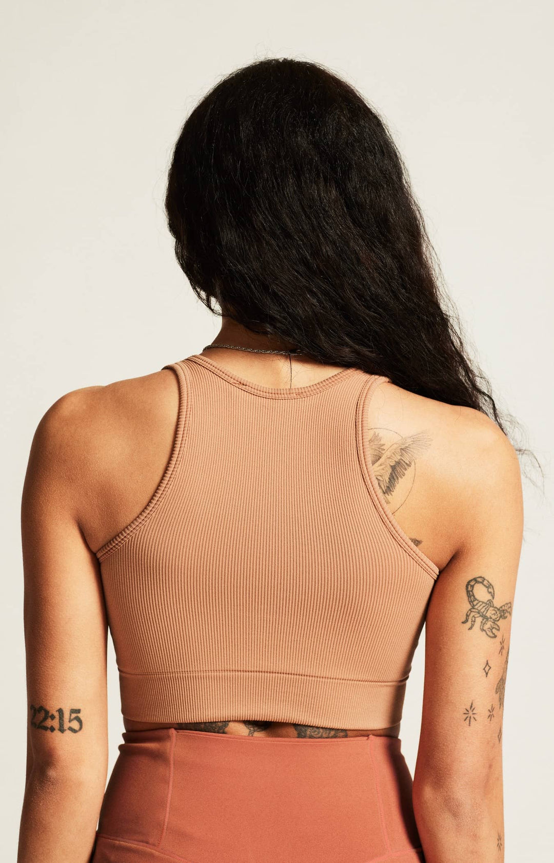 Person wearing a beige tank top with tattoos visible on arms and back against a plain background