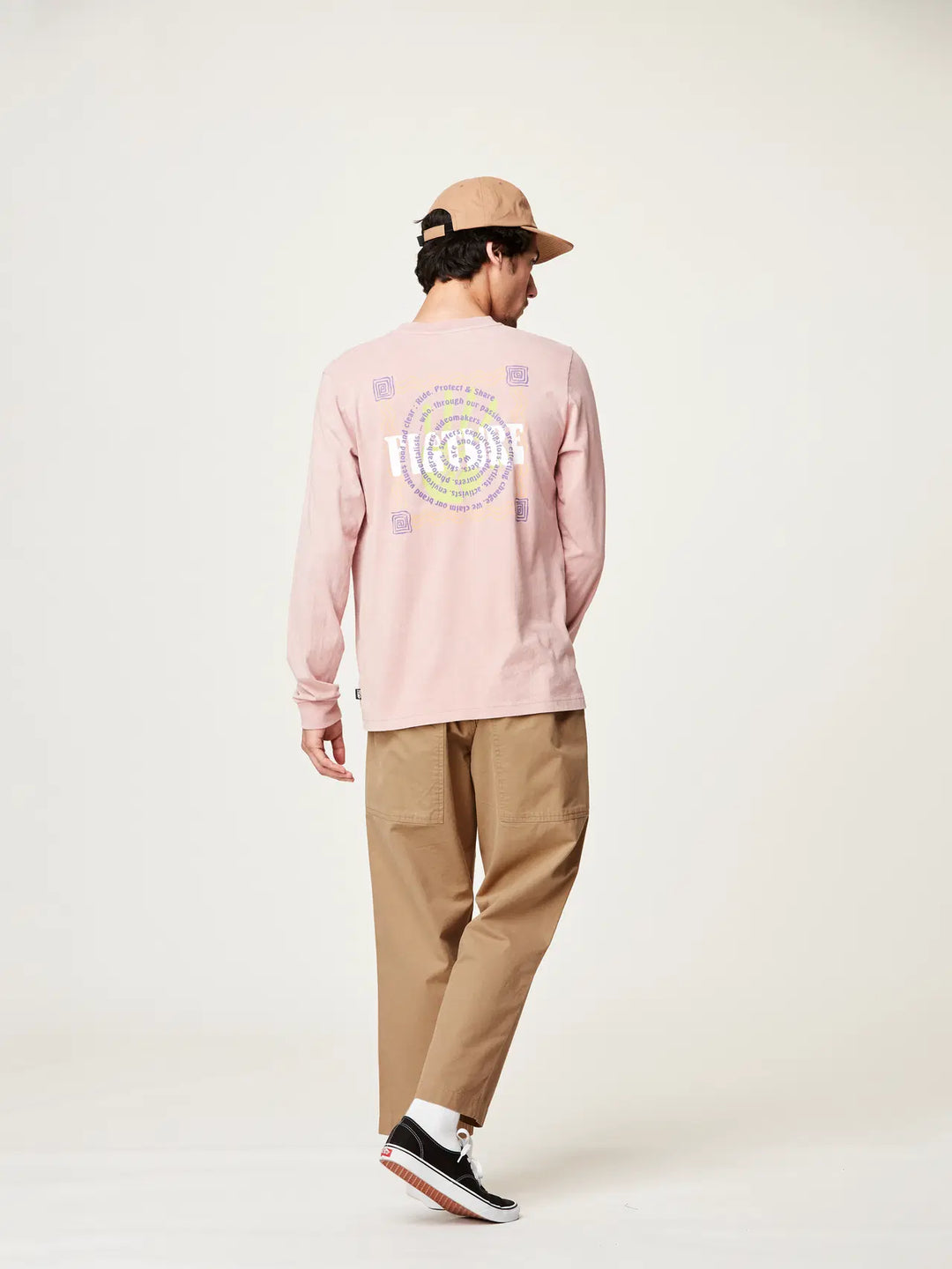 Person wearing a pink long-sleeve shirt with a logo, beige pants, and a cap on a white background