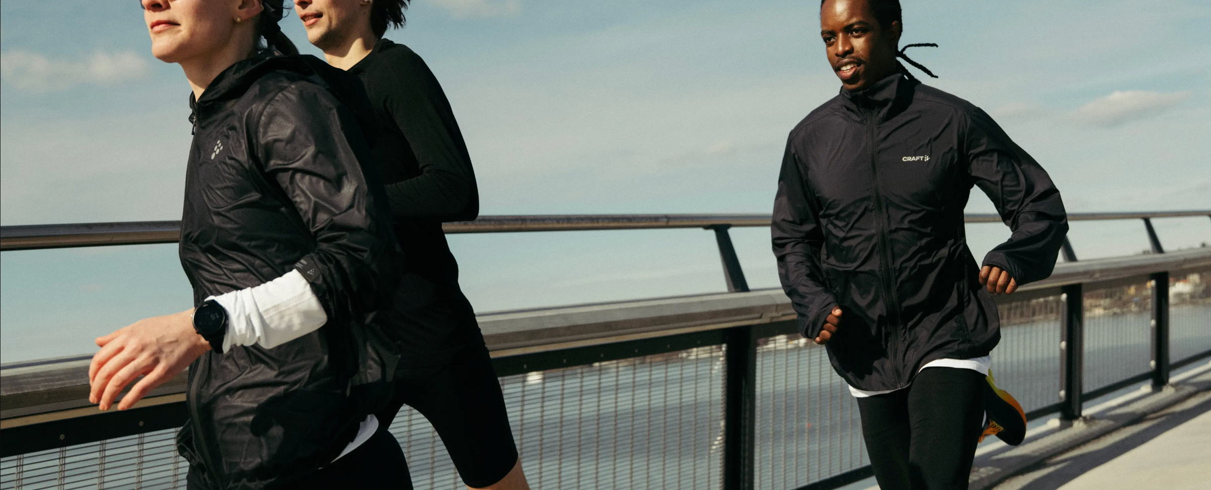 Three runners wearing black athletic wind jackets and tops jogging along an outdoor bridge under a cloudy sky.