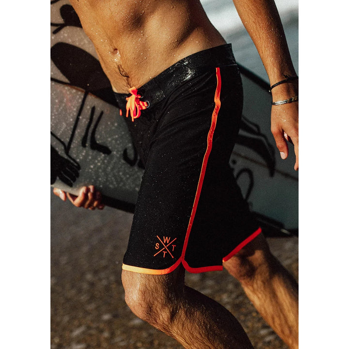 Person wearing black swim shorts with red accents and a logo, standing on a beach.